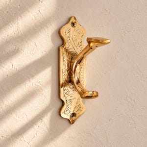May include: A polished gold-tone metal wall hook featuring an elongated, decorative backplate with a textured, stippled surface and engraved floral patterns. The hook has a curved, double-pronged design with a rounded top and bottom.