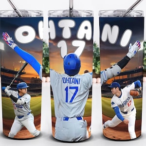 May include: A stainless steel tumbler featuring images of a baseball player in action. The tumbler has the name "Ohtani" and the number "17" printed on it. The background shows a baseball stadium at sunset. The tumbler includes a straw.