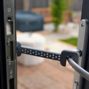May include: A gray door security bar is attached to a door frame. The bar has a series of small holes and connects to the door handle. The door frame and handle are silver.
