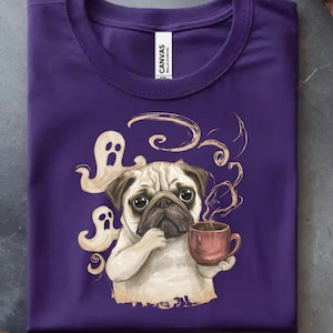 Könnte beinhalten: Ein violettes T-Shirt mit einer grafischen Illustration eines beigen Mopses, der eine braune Kaffeetasse hält, umgeben von zwei weißen Geisterfiguren und wirbelndem Dampf. Das T-Shirt hat einen Rundhalsausschnitt und ein weißes Etikett mit CANVAS.