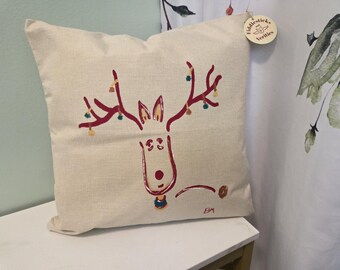 May include: A cream-colored square pillow with a whimsical reindeer design. The reindeer is depicted with maroon antlers adorned with colorful lights. The pillow has a tag that reads "Fiddlesticks and Verities".