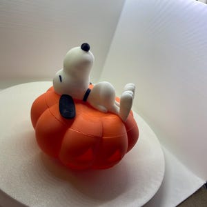 May include: A 3D printed figurine of a white beagle with a black ear and nose, lying on its back atop a bright orange pumpkin. The pumpkin features carved-out sections at the base, and the beagle has a black collar around its neck, resting peacefully.