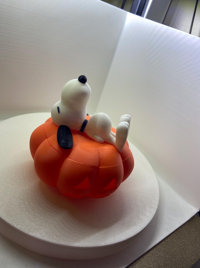 May include: A 3D printed figurine of a white beagle with a black ear and nose, lying on its back atop a bright orange pumpkin. The pumpkin features carved-out sections at the base, and the beagle has a black collar around its neck, resting peacefully.