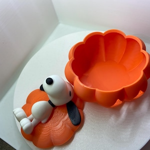 May include: A bright orange pumpkin-shaped container with a removable lid. The lid features a white cartoon dog figure with black ears and a black nose, lying flat on top. The container has a scalloped edge and a smooth, matte finish throughout.