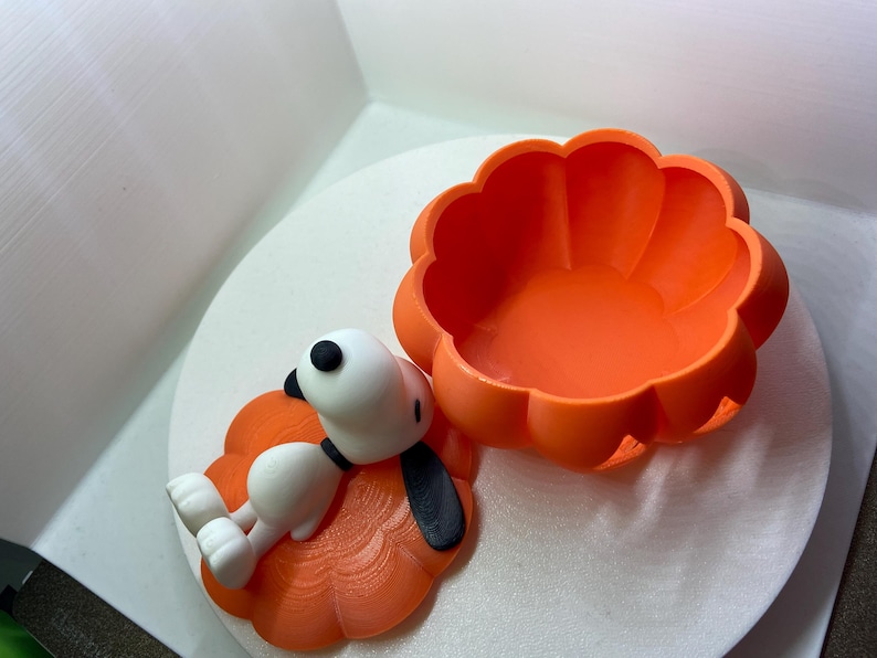 May include: A bright orange pumpkin-shaped container with a removable lid. The lid features a white cartoon dog figure with black ears and a black nose, lying flat on top. The container has a scalloped edge and a smooth, matte finish throughout.