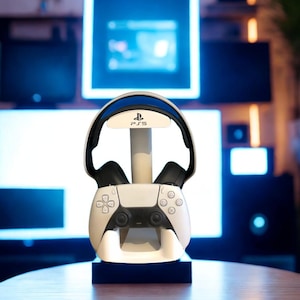 May include: A white gaming accessory stand featuring the PS5 logo, designed to hold a black and white wireless controller at the base and a pair of black over-ear gaming headphones on the top arch, providing a centralized storage solution.