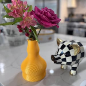 Puede incluir: Un jarrón amarillo brillante con una superficie texturizada de trama cruzada que contiene flores de alstroemeria rosas y una rosa rosa oscuro, junto a una pequeña figura de cerdo a cuadros blancos y negros con orejas y patas doradas.