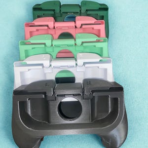 Può includere: Una pila di cinque impugnature per controller per un dispositivo di gioco portatile, disposte verticalmente. Le impugnature sono di colore verde scuro, rosa, verde chiaro, bianco e nero. Ogni impugnatura presenta marcature L e R e un foro.