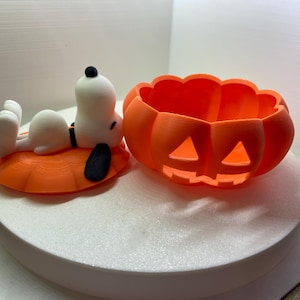 Könnte beinhalten: Eine weiße Snoopy-Figur mit schwarzen Ohren, die auf einem orangefarbenen, kürbisförmigen Deckel liegt, neben einer passenden orangefarbenen, kürbisförmigen Schale mit geschnitzten dreieckigen Augen und einem gezackten Mund, 3D-Druck-Optik.