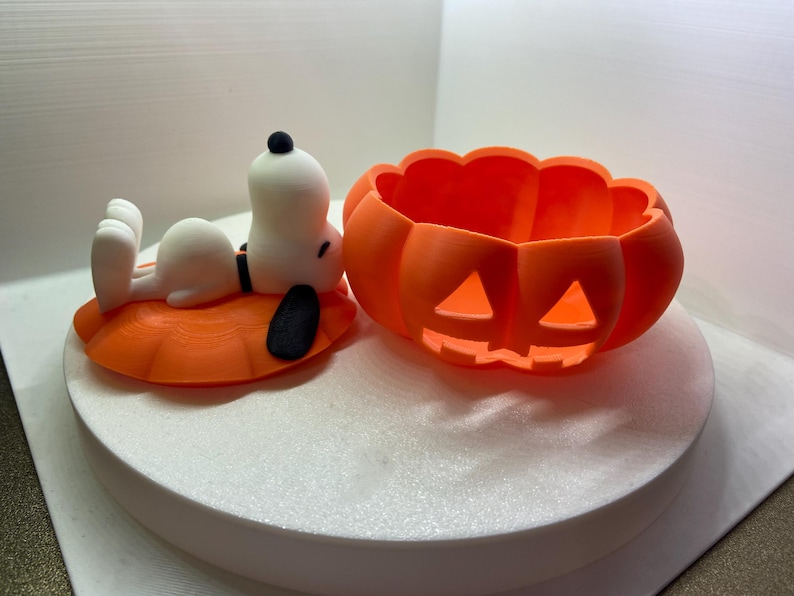 May include: A white Snoopy figure with black ears lying on an orange pumpkin-shaped lid next to a matching orange pumpkin-shaped bowl with carved triangular eyes and a jagged mouth, both featuring a textured, matte 3D-printed finish.