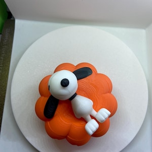 May include: A white and black cartoon dog figurine lying on top of a bright orange, multi-lobed pumpkin-shaped object. The dog has a round head, long black ears, and a small black nose, while the pumpkin base features a textured, layered surface.