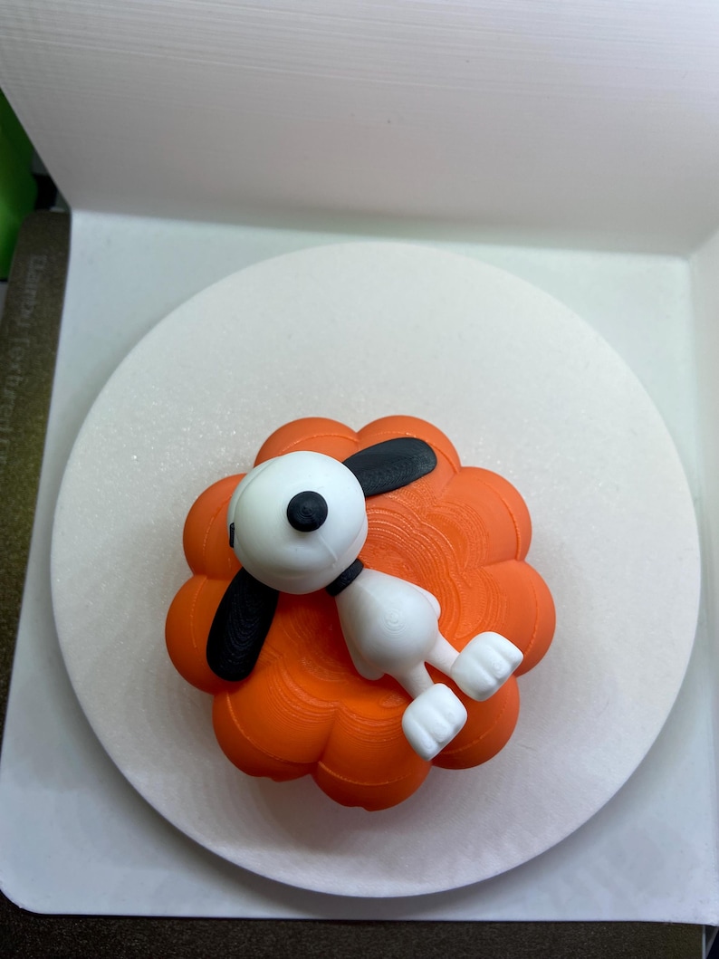 May include: A white and black cartoon dog figurine lying on top of a bright orange, multi-lobed pumpkin-shaped object. The dog has a round head, long black ears, and a small black nose, while the pumpkin base features a textured, layered surface.
