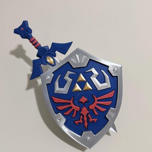 May include: A blue and silver shield featuring a central red bird emblem and three gold triangles, with a blue sword handle extending from the top. The shield has a silver border with small silver accents and a textured blue surface finish.