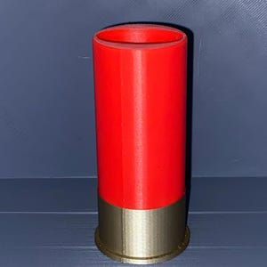 Pode incluir: Um recipiente cilíndrico concebido para se assemelhar a um cartucho de caçadeira, com uma secção superior vermelha brilhante e uma base de cor dourada metálica com uma borda. O objeto está na vertical e apresenta uma textura lisa e uniforme.
