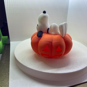May include: A white Snoopy figure with a black ear and collar resting on top of an orange jack-o'-lantern pumpkin. The pumpkin features carved triangular eyes and a jagged mouth opening, creating a hollow space within the orange plastic structure.