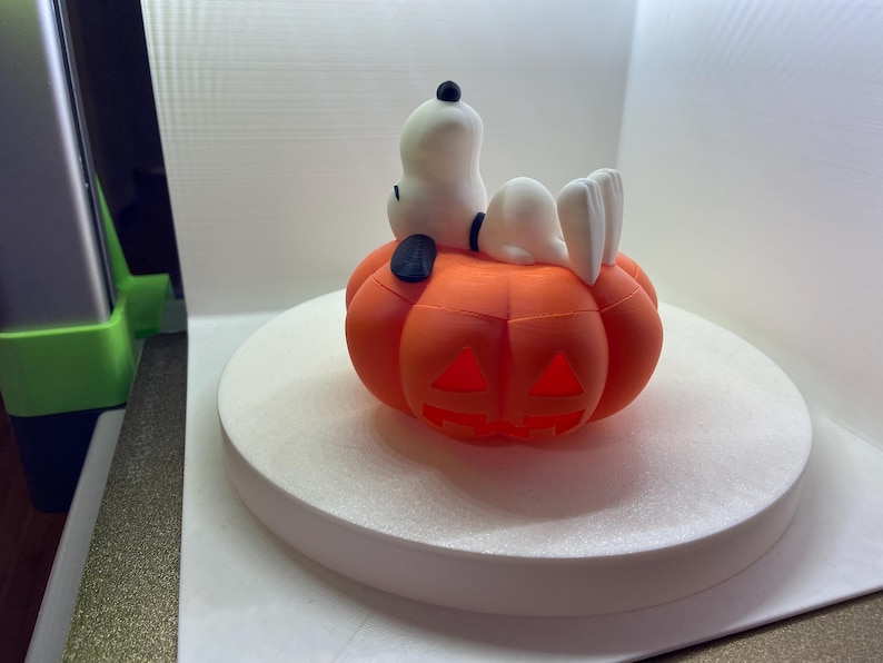 May include: A white Snoopy figure with a black ear and collar resting on top of an orange jack-o'-lantern pumpkin. The pumpkin features carved triangular eyes and a jagged mouth opening, creating a hollow space within the orange plastic structure.