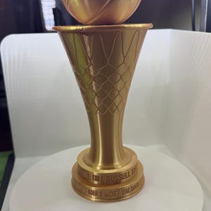 May include: A gold-colored basketball trophy featuring a spherical ball resting atop a flared base designed with a net pattern. The circular base includes embossed text reading THE BILL RUSSELL TROPHY and NBA FINALS MOST VALUABLE PLAYER.