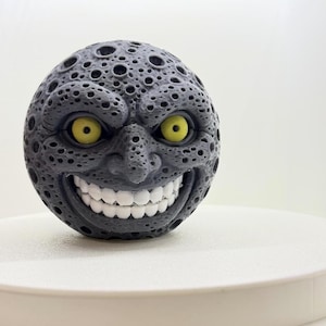 Peut inclure: Un objet sphérique avec une surface gris foncé parsemée de cratères rappelant la lune. Il présente un visage proéminent avec deux grands yeux jaunes, un nez et un large sourire montrant une rangée complète de dents blanches. La texture est poreuse.