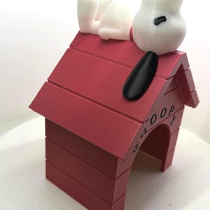 May include: A white cartoon dog figure with a black ear and nose resting on top of a red doghouse. The doghouse features a peaked roof and a rectangular opening at the front, with the name SNOOPY embossed in black lettering on the side of the roof.