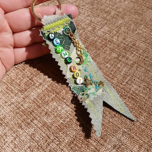 May include: A handmade keychain with a gold ring and a fabric tag. The tag is olive green with the words "LET ME ROT" spelled out in colorful beads. It features gold charms and decorative stitching.