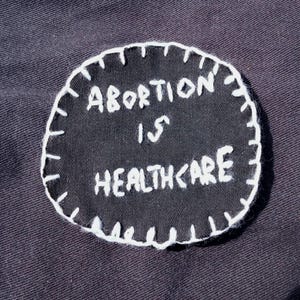 Puede incluir: Un parche negro circular con costuras blancas alrededor del borde. El parche presenta el texto bordado "ABORTION IS HEALTHCARE" en hilo blanco. El parche está sobre un fondo oscuro.