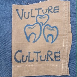 May include: A tan fabric patch with the words "VULTURE CULTURE" in dark blue, and a graphic of three teeth. The patch is sewn onto blue denim fabric.