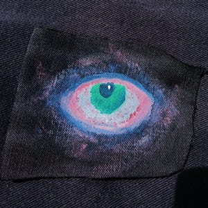 May include: A hand-painted patch featuring a stylized eye design. The eye has a green iris, pink and white details, and is surrounded by a blue and purple background, resembling a galaxy. The patch is on a dark denim fabric.