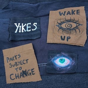 Op de afbeelding: Close-up van een donkerblauwe stof met vier patches. De patches hebben tekst en illustraties, waaronder de woorden "YIKES", "WAKE UP" en "PARTS SUBJECT TO CHANGE". E&eacute;n patch heeft een oogillustratie.
