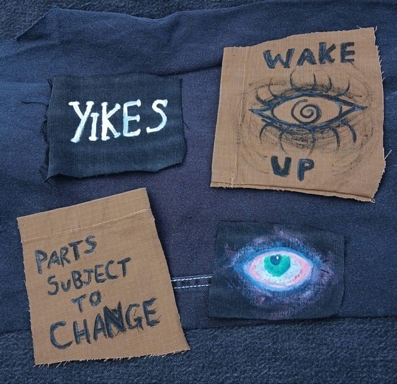 Op de afbeelding: Close-up van een donkerblauwe stof met vier patches. De patches hebben tekst en illustraties, waaronder de woorden "YIKES", "WAKE UP" en "PARTS SUBJECT TO CHANGE". E&eacute;n patch heeft een oogillustratie.