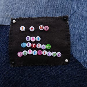 May include: A denim patch with the phrase "I ❤️ MY GAY LITTLE BOYFRIEND" spelled out in colourful letter beads. The patch is sewn onto a denim background with a patchwork design.