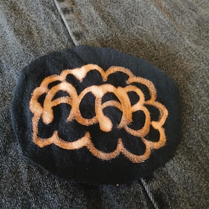 May include: A black, oval fabric patch featuring a light brown, abstract design. The patch is set against a denim background. The design is a stylized, interconnected pattern, suitable for clothing or accessories.