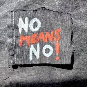 May include: A dark gray fabric patch with the words "NO MEANS NO!" in white and red lettering. The patch is sewn onto a darker gray fabric, possibly denim.