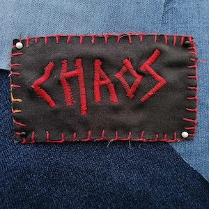 May include: A rectangular black patch with the red word "CHAOS" embroidered on it. The patch is stitched with red thread around the edges and is pinned to a denim background with white-headed pins. The denim background is a patchwork of different shades of blue.