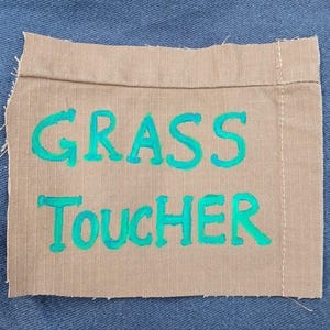 May include: A rectangular patch of tan fabric with the words "GRASS TOUCHER" in bright turquoise lettering. The patch is stitched onto a blue denim background, with visible stitching along the edges.