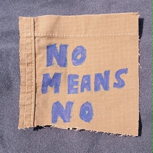 May include: A tan fabric patch with the words "NO MEANS NO" in blue paint. The patch has a stitched border and is set against a blue background.