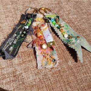 May include: Three handmade fabric keychains with metal rings. One is dark green with black accents and small beads. The second is colorful with a measuring tape detail. The third is light green with colorful beads and a peace sign charm.