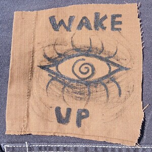 May include: A brown fabric patch with the words "WAKE UP" in black block letters. The patch features a stylized eye design in black, with a spiral pupil and radiating lines. The patch is sewn onto a blue denim fabric.