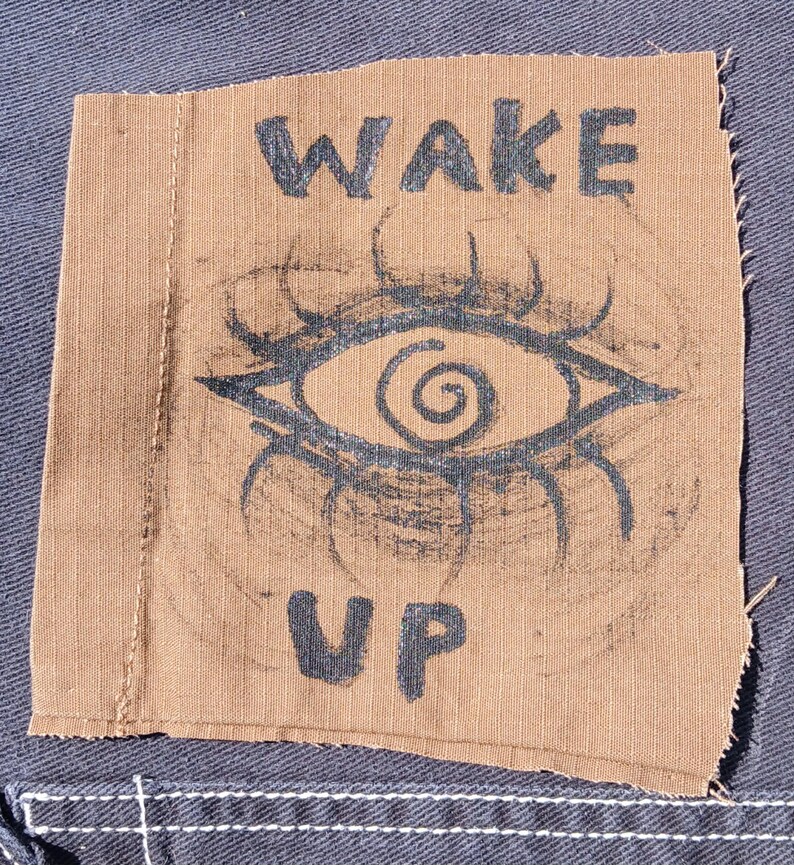 Op de afbeelding: Een bruine stoffen patch met de woorden "WAKE UP" in zwarte blokletters. De patch heeft een gestileerd oogontwerp in zwart, met een spiraalvormige pupil en stralende lijnen. De patch is op een blauwe denim stof genaaid.