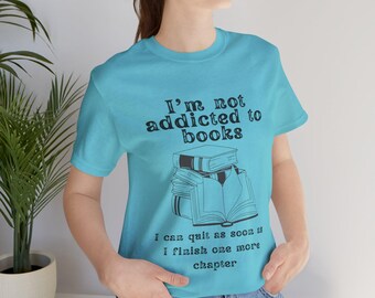 Könnte beinhalten: Ein türkisfarbenes T-Shirt mit dem Text "I'm not addicted to books" und einem Stapel Bücher mit einem offenen Buch obenauf. Der Text unter den Büchern lautet "I can quit as soon as I finish one more chapter."