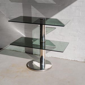 Peut inclure: Une table d'appoint à deux niveaux dotée d'une base circulaire en chrome poli et d'une colonne de support centrale verticale en chrome. La table possède deux étagères rectangulaires en verre teinté foncé et un capuchon chromé sur le dessus.