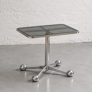 Puede incluir: Una mesa auxiliar rectangular con un tablero de cristal ahumado oscuro de esquinas redondeadas y una estructura de cromo pulido. La base consta de una columna central vertical apoyada por cuatro patas radiantes, cada una con un pie esférico.