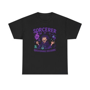 Op de afbeelding: Zwart t-shirt met een cartoon tovenaar ontwerp in paars. De tovenaar houdt een gloeiende bol en een drankfles vast, met de tekst "SORCERER OF QUESTIONABLE DECISIONS". Een kikker is ook aanwezig in het ontwerp.