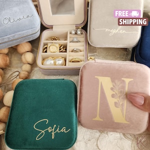 May include: A collection of square velvet jewellery travel cases in various colours including light blue, beige, dark green, and dusty pink. Each case features a gold zip and personalised text or initials. One open case displays rings and earrings.