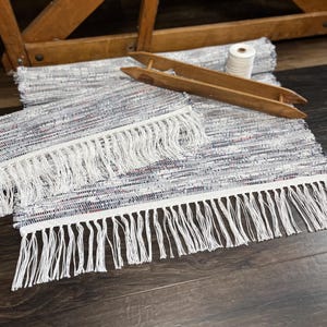 May include: Handwoven rag rug with white fringe. The rug features a mix of blue, red, and white fabric. A wooden shuttle and a spool of white thread are also visible. The rug is likely handmade and suitable for home decor.