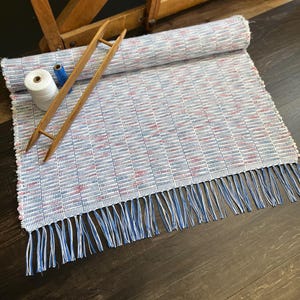 May include: A handwoven rug with a blue, white, and red checkered pattern, featuring a fringe of blue and white threads. A wooden shuttle and spools of white and blue thread are placed on the rug, suggesting a craft or textile theme.