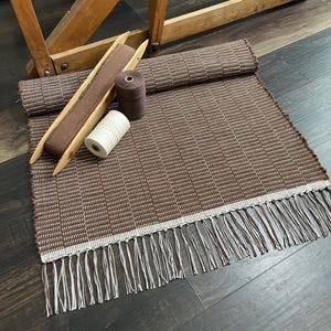 May include: A woven brown and white rug with a fringed edge. A wooden shuttle with brown fabric, two spools of thread, and a wooden loom are also in the image. The rug is rectangular and has a woven pattern.