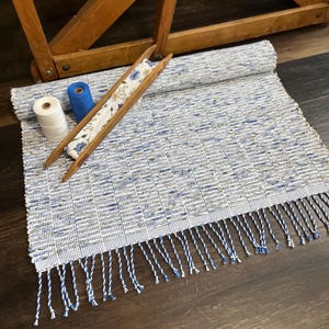 May include: A woven rug with a blue and white pattern, featuring a fringe along one edge. A wooden shuttle with floral fabric and two spools of thread are on the rug. The rug is rolled up in the background.
