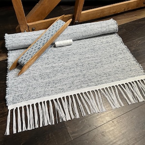 May include: A rolled-up gray and white woven rug with a white fringe. A wooden shuttle with a blue and white geometric pattern is placed on the rug, along with a spool of white thread. The rug is on a dark wood surface.