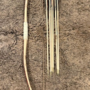 May include: A traditional wooden bow with a red and black accent, and a set of arrows. The bow is wrapped with white material at the grip. The arrows have black and red fletching. The bow and arrows are set against a textured brown background.