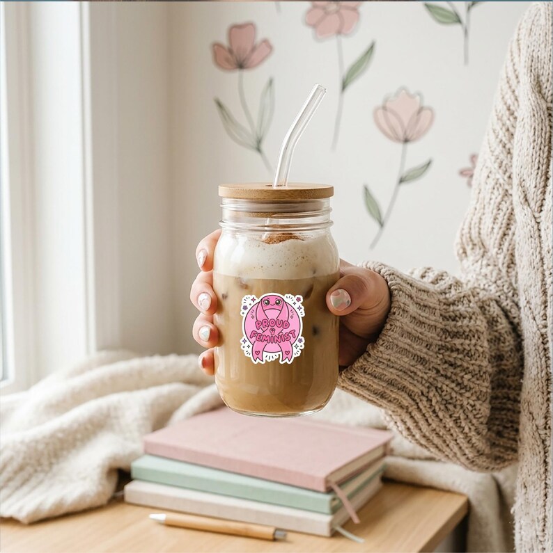 Pode incluir: Um frasco de vidro transparente com tampa de madeira e canudo transparente, cheio de uma bebida de caf&eacute;. Um adesivo rosa com as palavras "Proud Feminist" est&aacute; no frasco. O frasco &eacute; segurado em frente a uma pilha de livros.